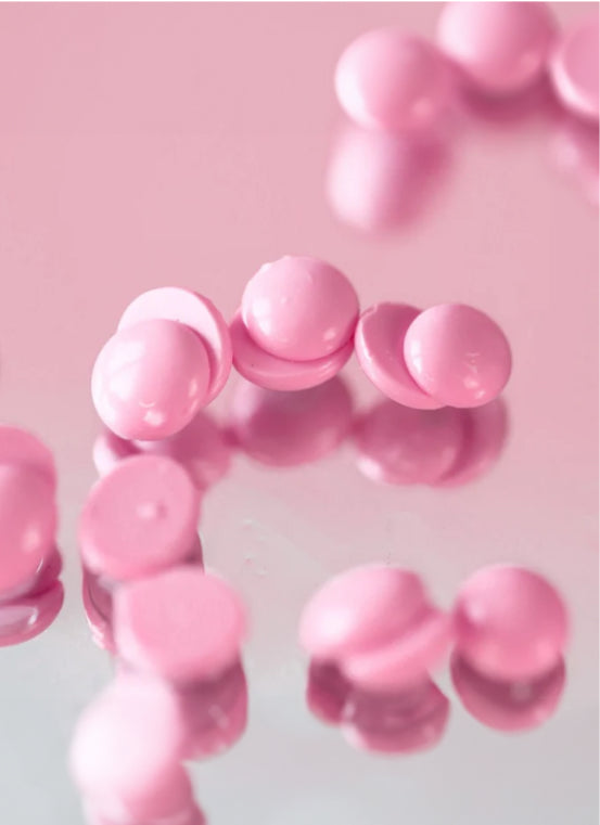Close-up of scattered pink round pills on a reflective pink surface, creating soft reflections. The pills are shiny and uniformly shaped, with shadows adding depth to the image.