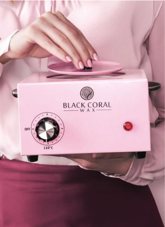 Person adjusting the lid of a pink Black Coral Wax warmer, with a temperature dial and power switch on the front. The person is wearing a light pink blouse and maroon skirt.