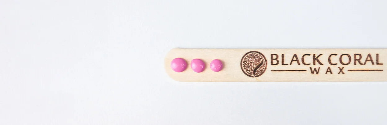 A wooden spatula with BLACK CORAL WAX and a logo engraved on it. Three small pink wax beads are placed on the left side of the spatula against a plain white background.