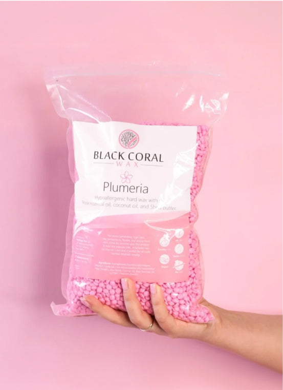 A hand holds a clear bag filled with small pink wax beads labeled Black Coral Wax Plumeria. The packaging highlights its hypoallergenic properties and ingredients like rosehip oil, coconut oil, and shea butter. The background is a soft pink color.