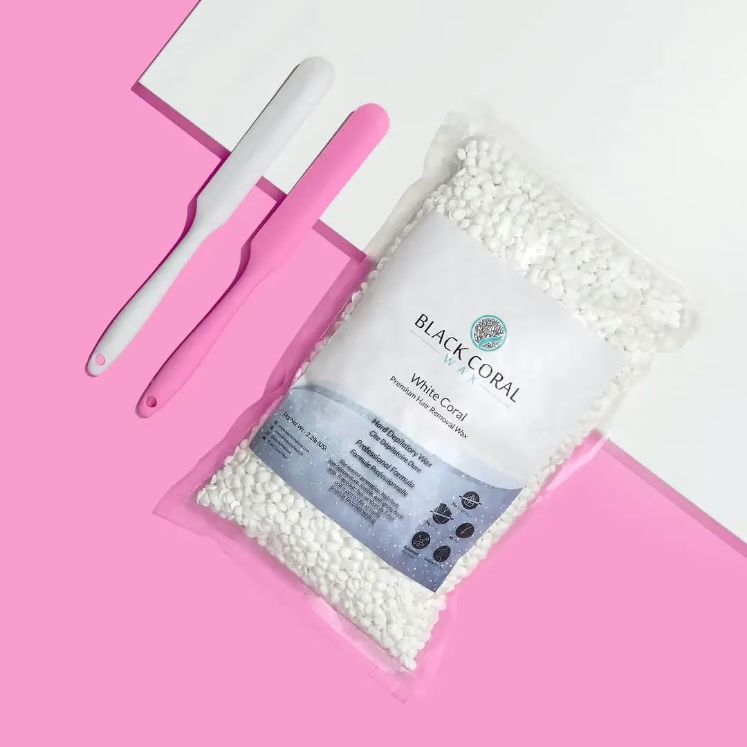 A 5lb Warmer + FREE Hard Wax from Black Coral Wax USA, filled with white wax beads, is shown on a pink and white background beside two waxing spatulas—one white and one pink.
