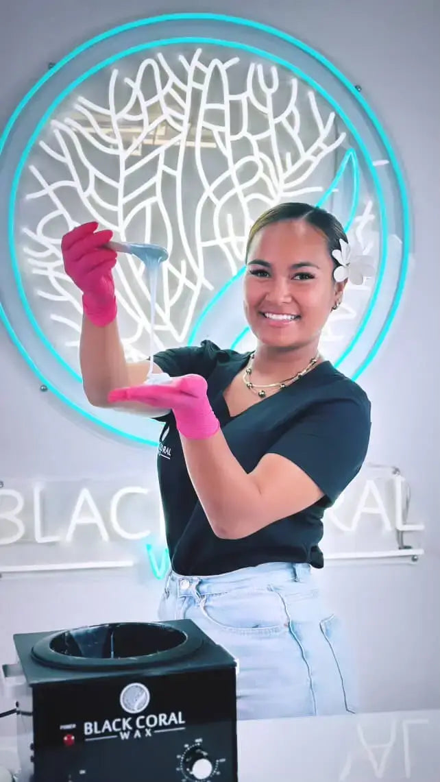 A smiling person with a flower in their hair holds a wax strip and wears pink gloves. They stand in front of a circular neon sign featuring a coral design and a Black Coral Wax sign. They are dressed in a black shirt and light blue jeans.