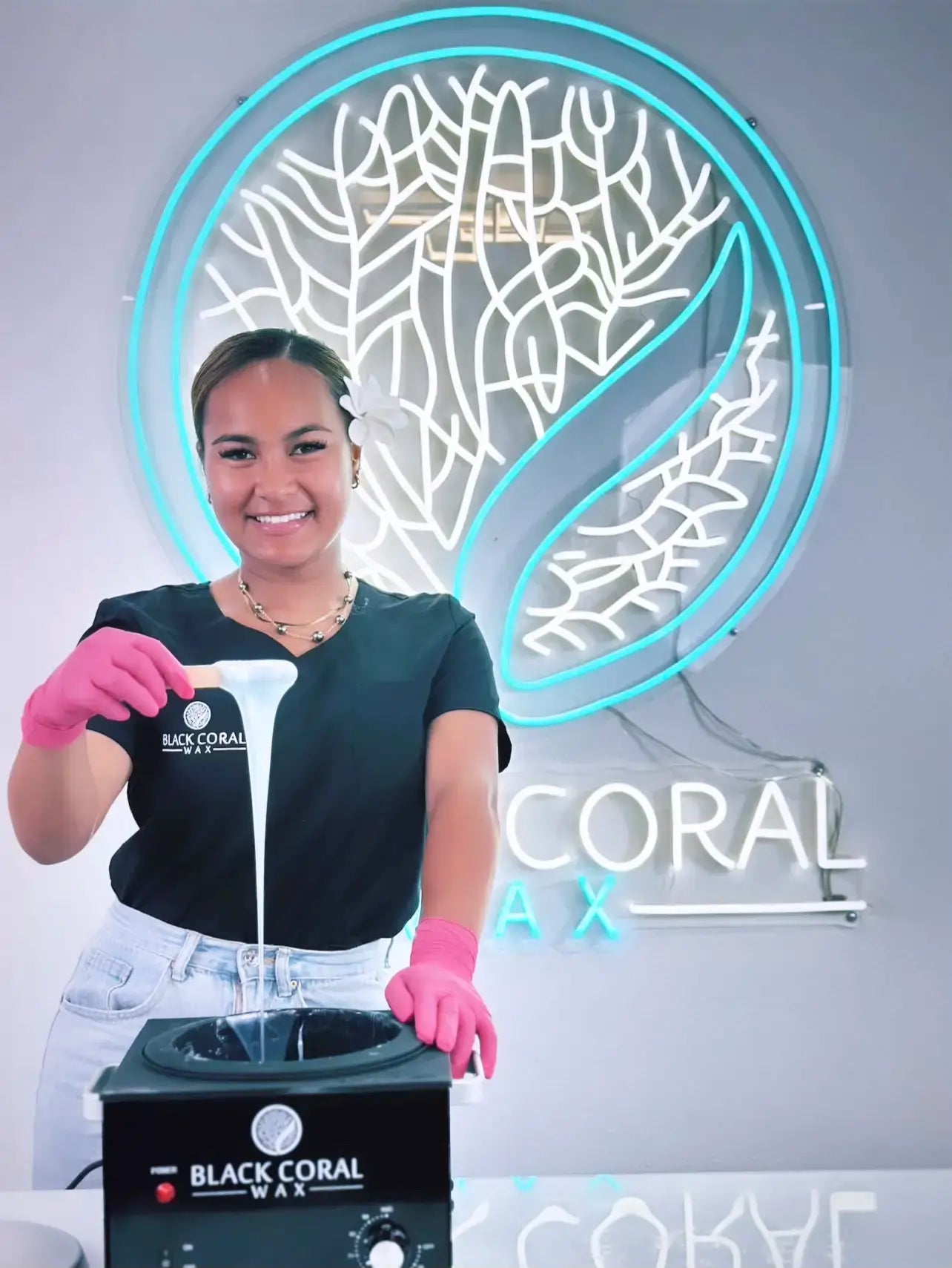 A person wearing a black t-shirt and pink gloves holds a spatula with wax dripping from it. Behind them is a neon sign featuring a stylized leaf logo and the words Black Coral Wax in white and blue.