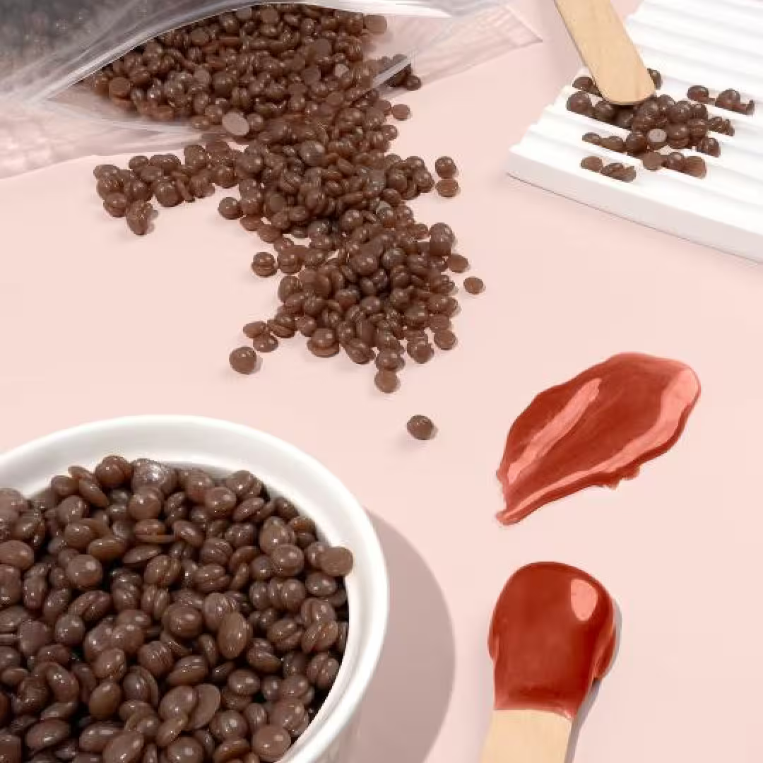 Chocolate-coated candies are spilled from a bag onto a pink surface. Nearby, two puddles of melted Pele Hard Wax Beads by Black Coral Wax USA with wooden sticks create the illusion of sweet treats or popsicles.