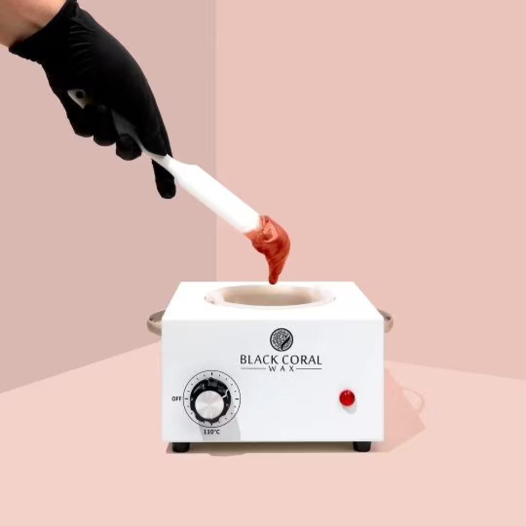 A hand in a black glove holds a spatula with melted Pele Hard Wax Beads above a white wax warmer labeled Black Coral Wax USA, set against a pink and beige background—ideal for removing coarse hair.