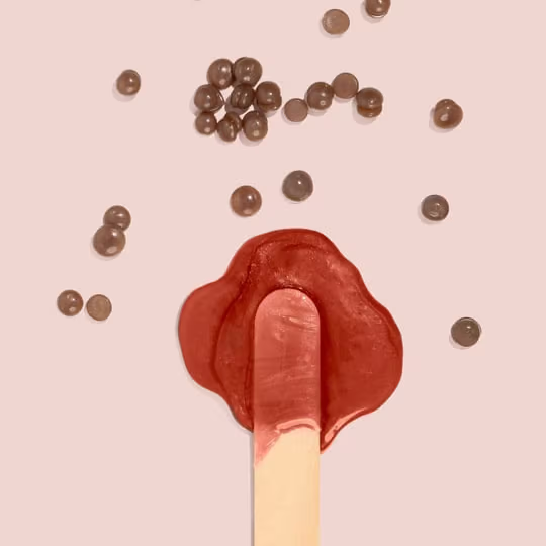 A melting red popsicle on a light pink background, surrounded by small round chocolate candies and Pele Hard Wax Beads from Black Coral Wax USA scattered above and around it.