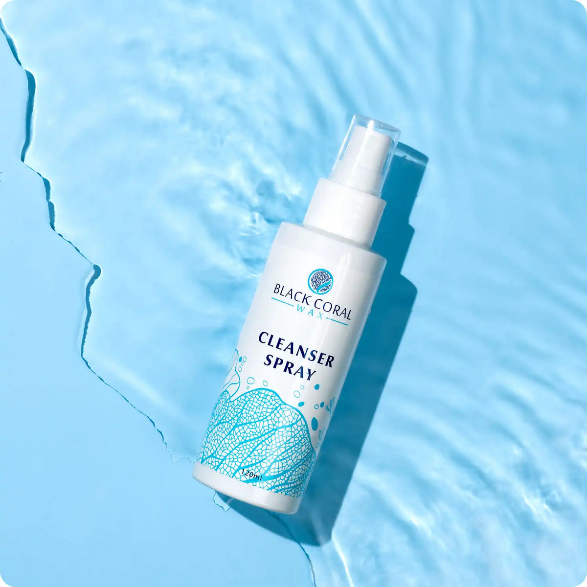 A bottle of Cleanser Spray 400ml from Black Coral Wax USA, a gentle pre-waxing cleanser, rests on a light blue surface near water, featuring seaweed designs on the label and rippling reflections beside it.
