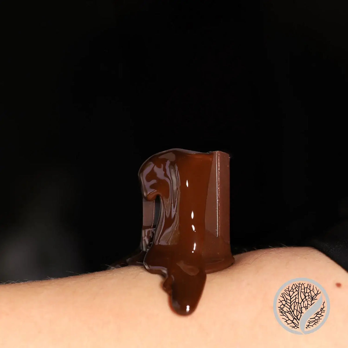 A piece of chocolate with melted chocolate drips on a light-skinned arm, evoking the smooth texture of Chocolate Hard Wax 400g by Black Coral Wax USA. A small tree-like logo appears in the bottom right corner on a black background.