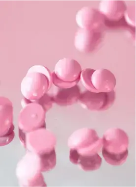 Pink round pills scattered on a reflective surface, creating soft reflections. The background is a matching light pink color.