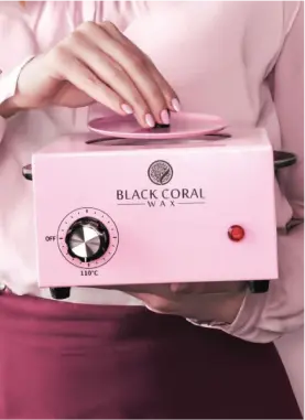 A person wearing a pink shirt holds a pink wax warmer with the brand Black Coral Wax on the front. The warmer has a temperature dial and a red indicator light. The lid is being lifted slightly.