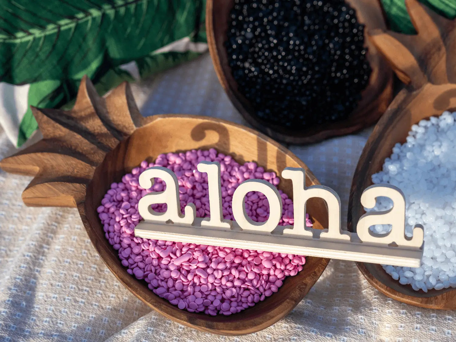 Wooden bowls shaped like pineapples contain small pellets in black, white, and pink. In the pink pellets, a wooden cutout spells aloha. The background features a fabric with a green leafy pattern.
