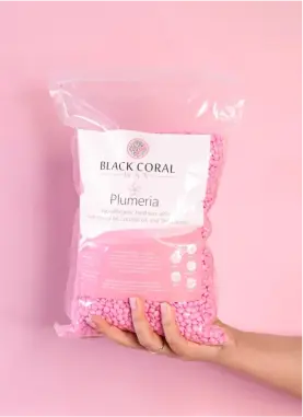 A hand holding a large, clear plastic bag filled with small pink beads against a pink background. The bag is labeled Black Coral Hawaii and Plumeria.