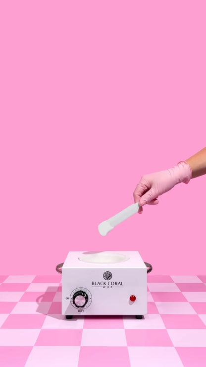 A gloved hand holds a spatula above a wax warmer filled with White Coral Wax by Black Coral Wax USA, ideal for gentle hair removal on sensitive skin, set on a pink checkered surface against a solid pink background.