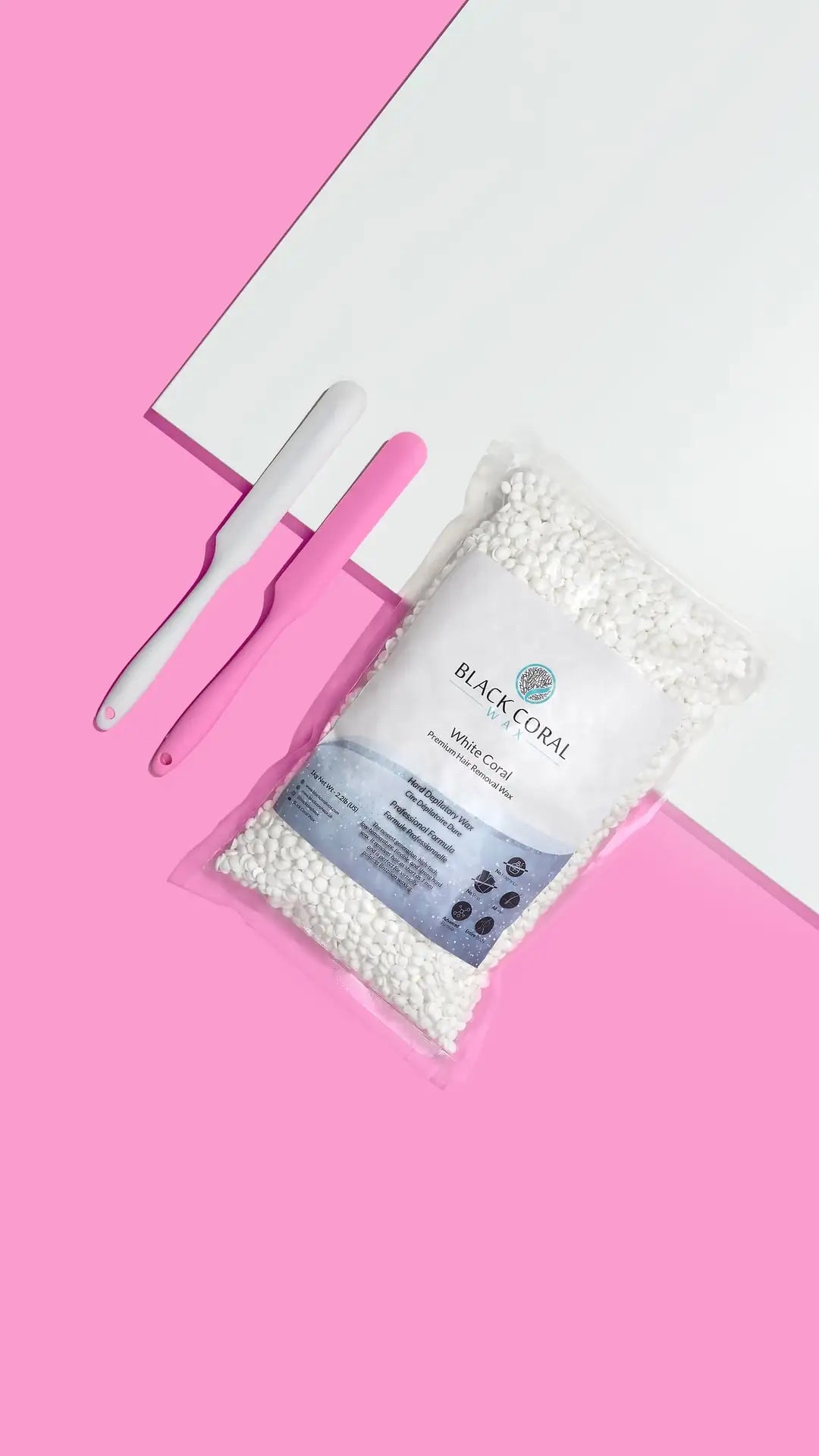 A bag of White Coral Wax by Black Coral Wax USA and two waxing spatulas—one white with a pink handle, one all pink—on a pink and white background, ideal for gentle hair removal on sensitive skin.
