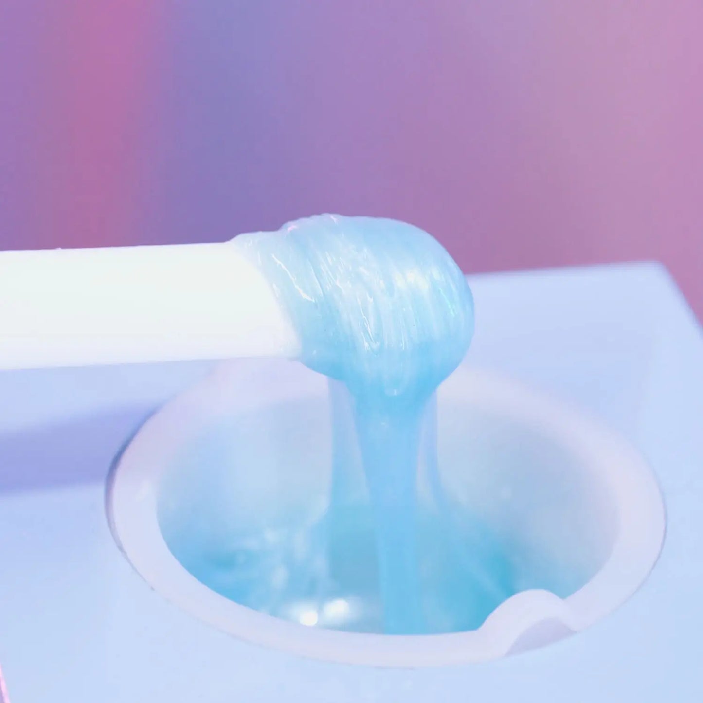 A close-up of Black Coral Wax USAs Kai Hard Wax, translucent blue and ideal for sensitive skin, being dispensed from a white tube into a round container against a softly blurred pink and purple backdrop.