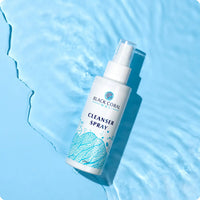 A bottle of Cleanser Spray 400ml from Black Coral Wax USA, a gentle pre-waxing cleanser, rests on a light blue surface near water, featuring seaweed designs on the label and rippling reflections beside it.