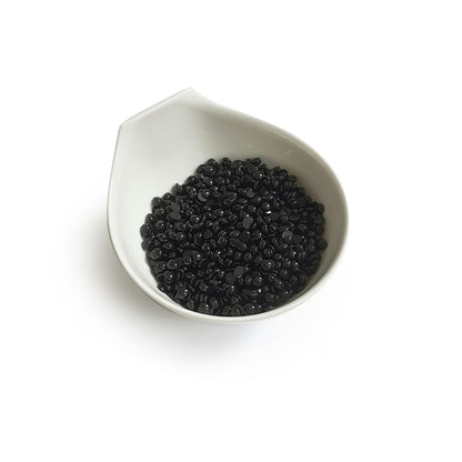 Black Coral Hard Wax  by Black Coral Wax USA is showcased in a white ceramic bowl akin to a Black Coral Hard Wax vessel. The bowls subtle, spout-like design elegantly cradles small, shiny black beads against a plain backdrop, reflecting potential for long-lasting hair removal.
