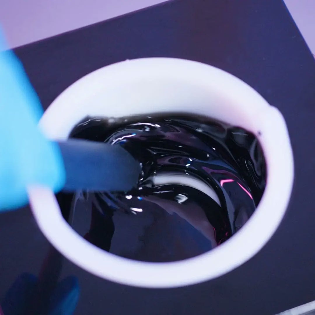 A person wearing a blue glove stirs a bowl of glossy Black Coral Hard Wax by Black Coral Wax USA with a black spatula. The thick, shiny liquid swirls and reflects light, against a dark, blurred background. This stripless wax shines with potential for long-lasting hair removal success.
