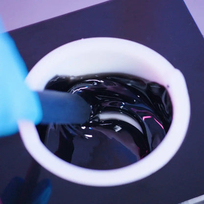 A person wearing a blue glove stirs a bowl of glossy Black Coral Hard Wax by Black Coral Wax USA with a black spatula. The thick, shiny liquid swirls and reflects light, against a dark, blurred background. This stripless wax shines with potential for long-lasting hair removal success.