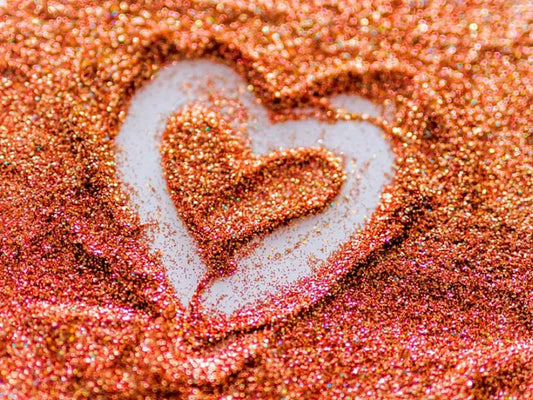 A heart shape is crafted with scattered, shimmering Boss Babe Glitter by Black Coral Wax USA on a surface, revealing a lighter background in the less-covered area. The orange and gold glitter sparkles brilliantly under the light.