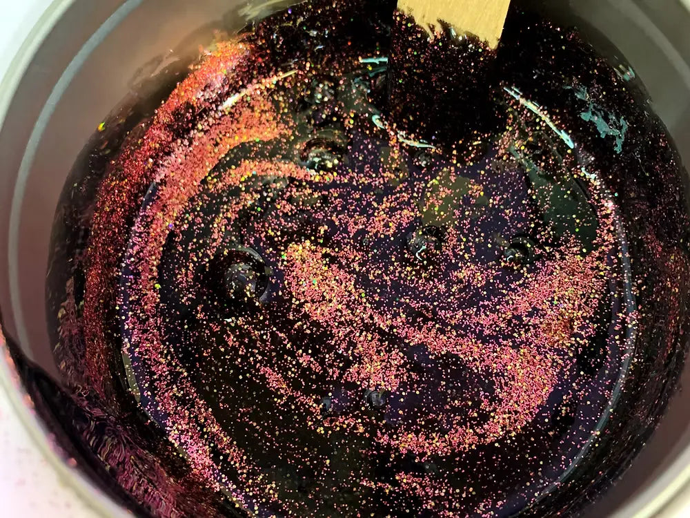 A pot of dark purple liquid swirling with Boss Babe Glitter Wax Creations from Black Coral Wax USA, stirred with a wooden stick, creates a shimmering galaxy-like effect with gold flecks, embodying dazzling artistry.