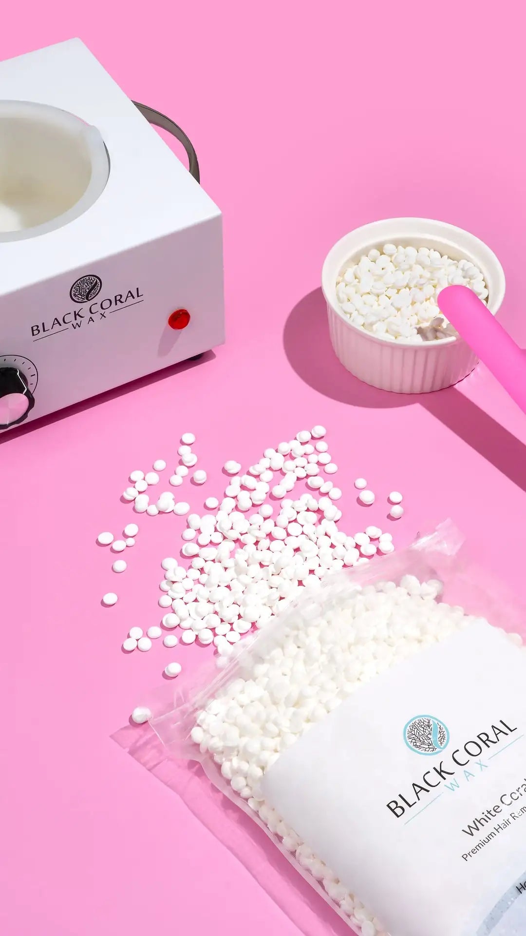 The White Coral Wax by Black Coral Wax USA includes a white wax warmer, small bowl, and open bag of hard wax beads on a pink surface with scattered beads and a pink spatula—ideal for gentle hair removal on sensitive skin.