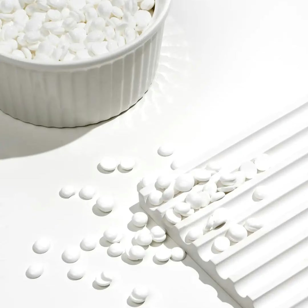White Coral Wax by Black Coral Wax USA—small white round tablets for hair removal—are scattered on a white surface near a fluted dish, creating a minimal scene that suggests gentle, sensitive skin care.