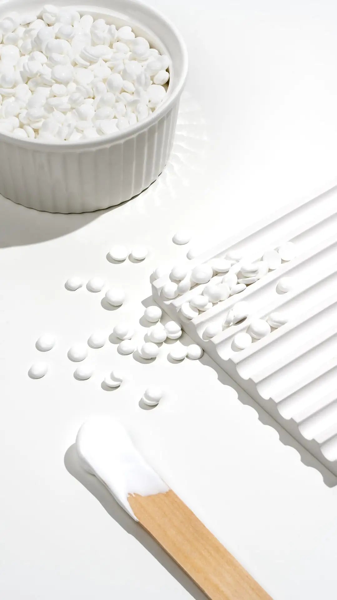 A bowl of White Coral Wax hard wax pellets by Black Coral Wax USA, scattered on a white grooved tray, with a spatula of creamy wax for hair removal—all arranged on a bright white surface.