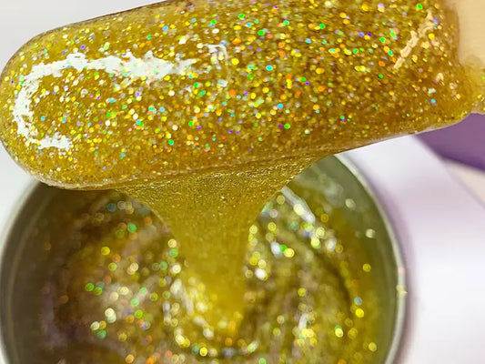 A close-up shows a wooden stick lifting thick, shimmering gold gel from a Dripping in Gold Glitter 2oz container by Black Coral Wax USA. The vibrant glitter wax sparkles with various reflections, creating a dazzling effect as the container is mostly filled with luxurious gel.