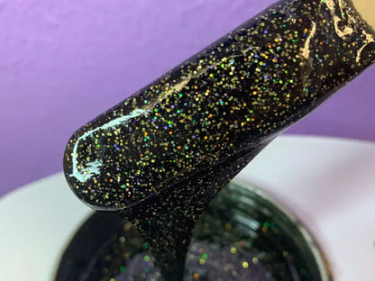 A close-up of a brush dipped in Black Coral Wax USAs Dripping in Gold Glitter 2oz from Glitter Wax Creations™ reveals multicolored and gold glitter nail polish. The violet background highlights the glossy, textured finish, turning the bottle into a shimmering masterpiece.