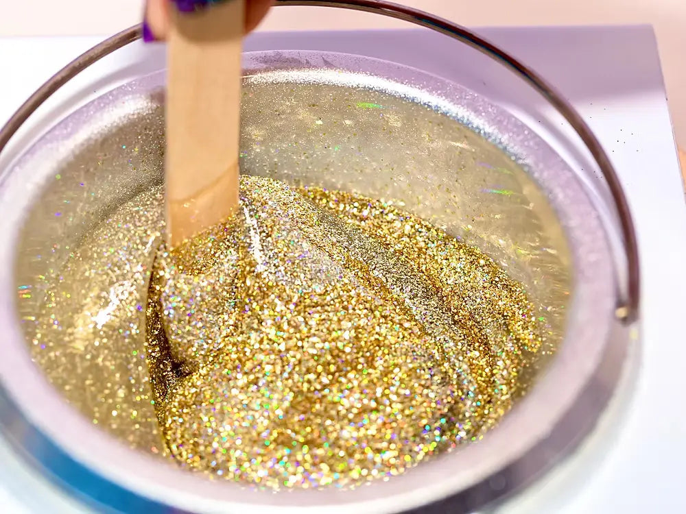 A hand stirs a shimmering substance in a metallic bowl using a wooden stick. The Dripping in Gold Glitter 2oz Glitter Wax Creations™ by Black Coral Wax USA reflects light with sparkling colors.