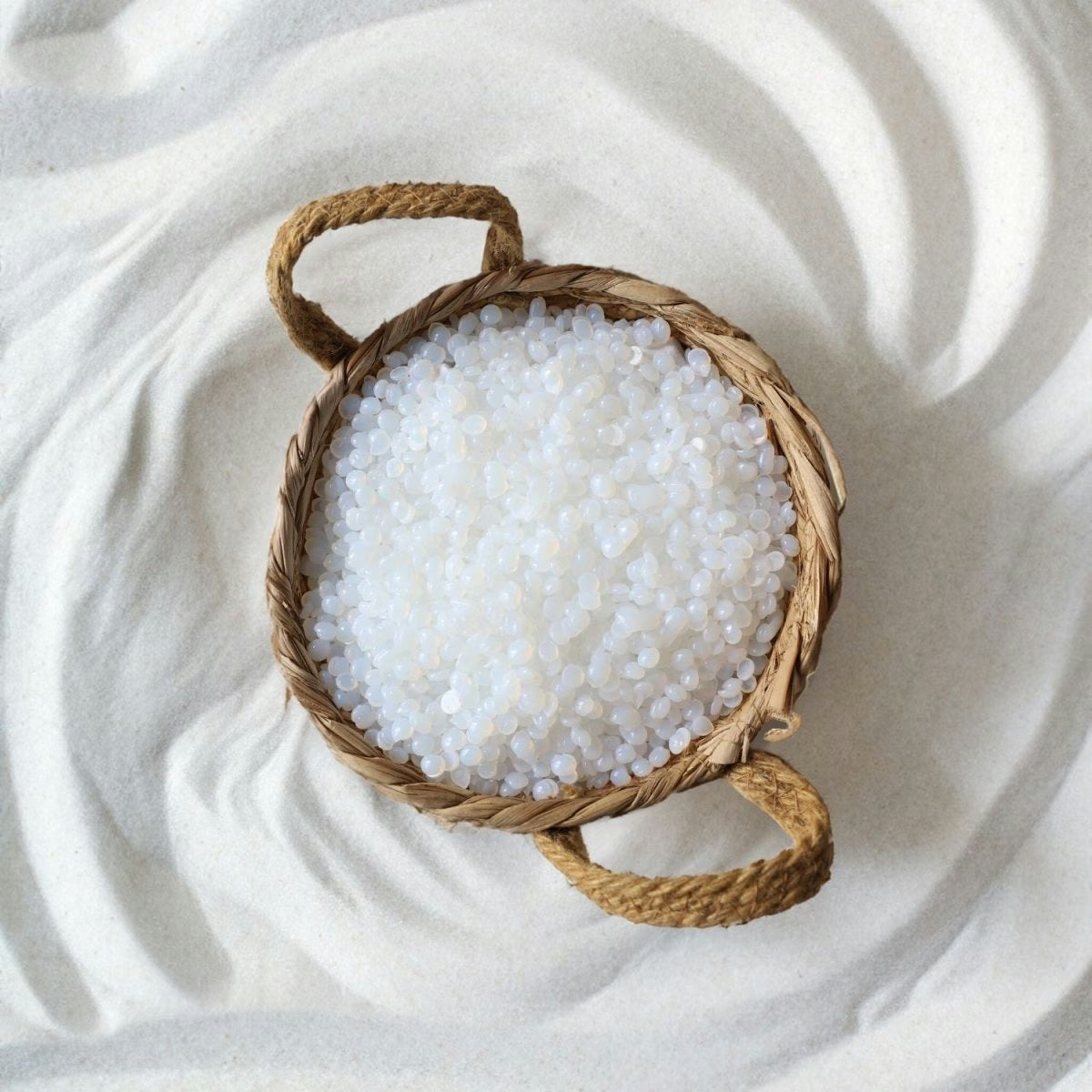 A round wicker basket with two handles holds Black Coral Wax USA’s Mae Mae Waxing Kit—5 bags of hard wax and cosmetics—resting on a wavy, textured white sand surface.