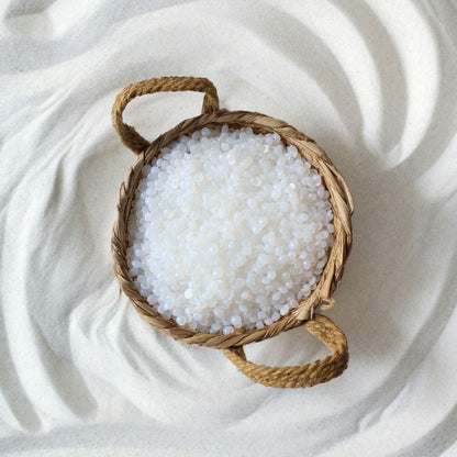 A round wicker basket with two handles holds Black Coral Wax USA’s Mae Mae Waxing Kit—5 bags of hard wax and cosmetics—resting on a wavy, textured white sand surface.