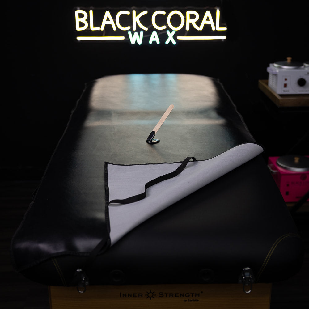 In a dimly lit room, a Waxing Pad by Black Coral Wax USA is draped with a partially pulled-back white sheet. A waxing spatula and small wax cup sit on the faux black leather. Nearby, two wax heaters stand under a neon BLACK CORAL WAX sign.