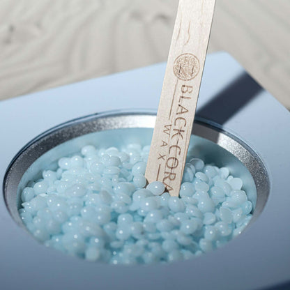 A close-up of a container filled with light blue Kai Hard Wax beads and a wooden stick engraved with Black Coral Wax USA. Ideal for sensitive skin, the stick is partially submerged in the beads against a softly blurred background.
