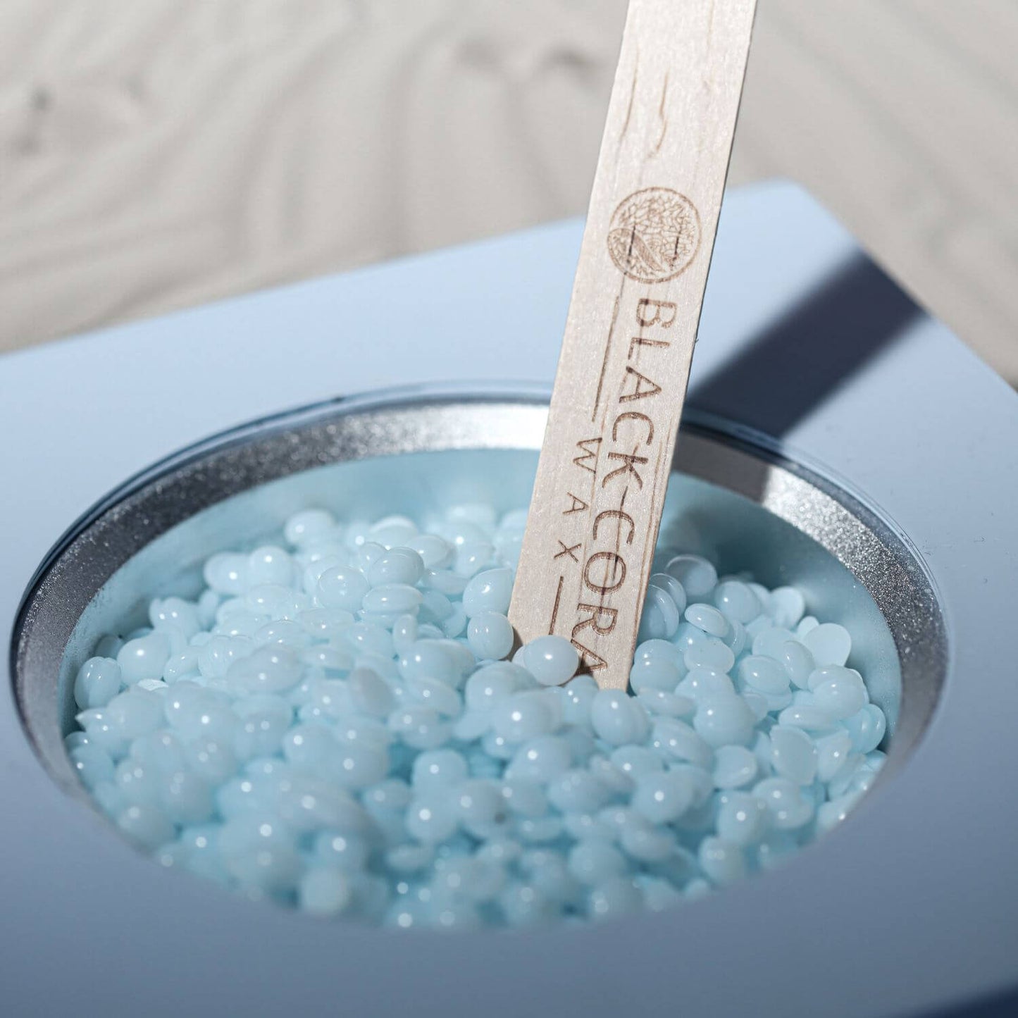 A close-up of a container filled with light blue Kai Hard Wax beads and a wooden stick engraved with Black Coral Wax USA. Ideal for sensitive skin, the stick is partially submerged in the beads against a softly blurred background.