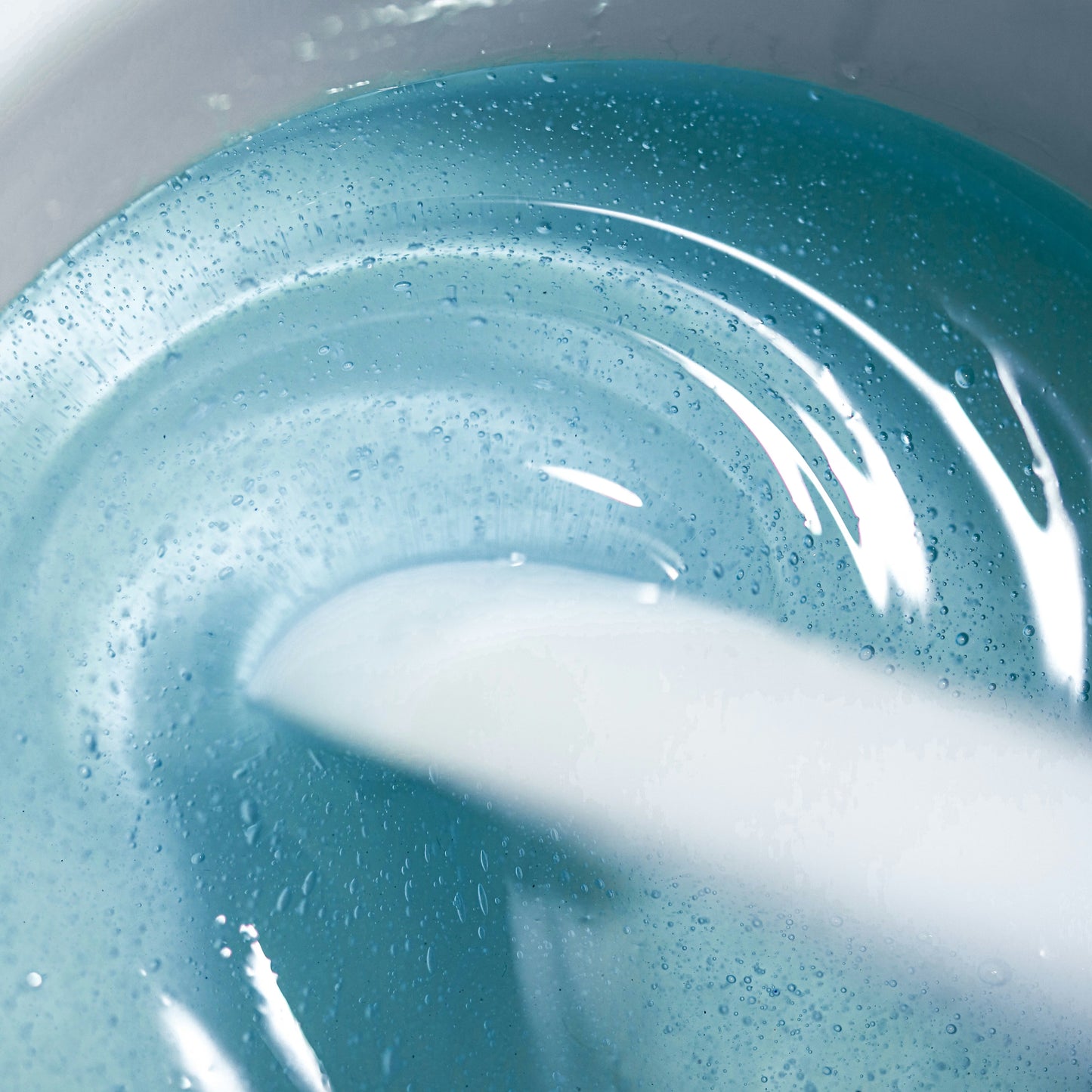 In a white container, the Kai Hard Wax by Black Coral Wax USA is shown as it swirls in blue liquid, creating a glossy sheen with light reflections. A partially submerged white object stirs the wax for hair removal on sensitive skin, forming small bubbles.
