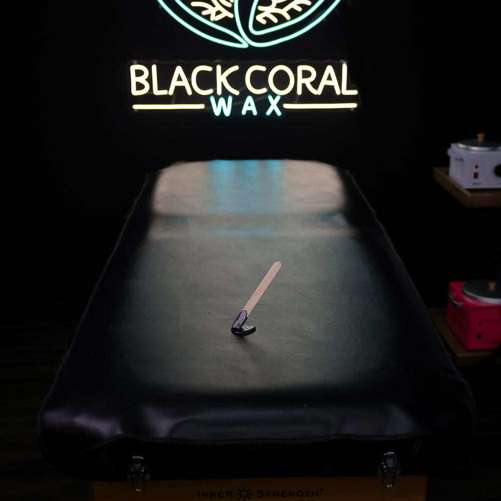 A dark Waxing Pad covered in faux black leather holds a spatula and wax. In the background, a neon sign with a leaf design says BLACK CORAL WAX USA, casting a gentle glow.