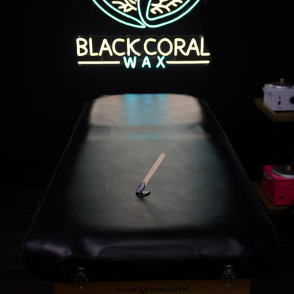 A dark Waxing Pad covered in faux black leather holds a spatula and wax. In the background, a neon sign with a leaf design says BLACK CORAL WAX USA, casting a gentle glow.