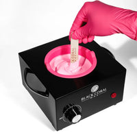A gloved hand dips a wooden spatula into pink hair removal wax in the Single (1lb) Wax Warmer by Black Coral Wax USA, featuring a dial and red power button, shown on a white background.