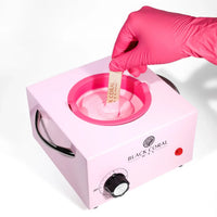 A hand in a pink glove stirs melted wax with a spatula in the Black Coral Wax USA Single (1lb) Wax Warmer, which features a temperature dial and red power button—ideal for efficient hair removal.