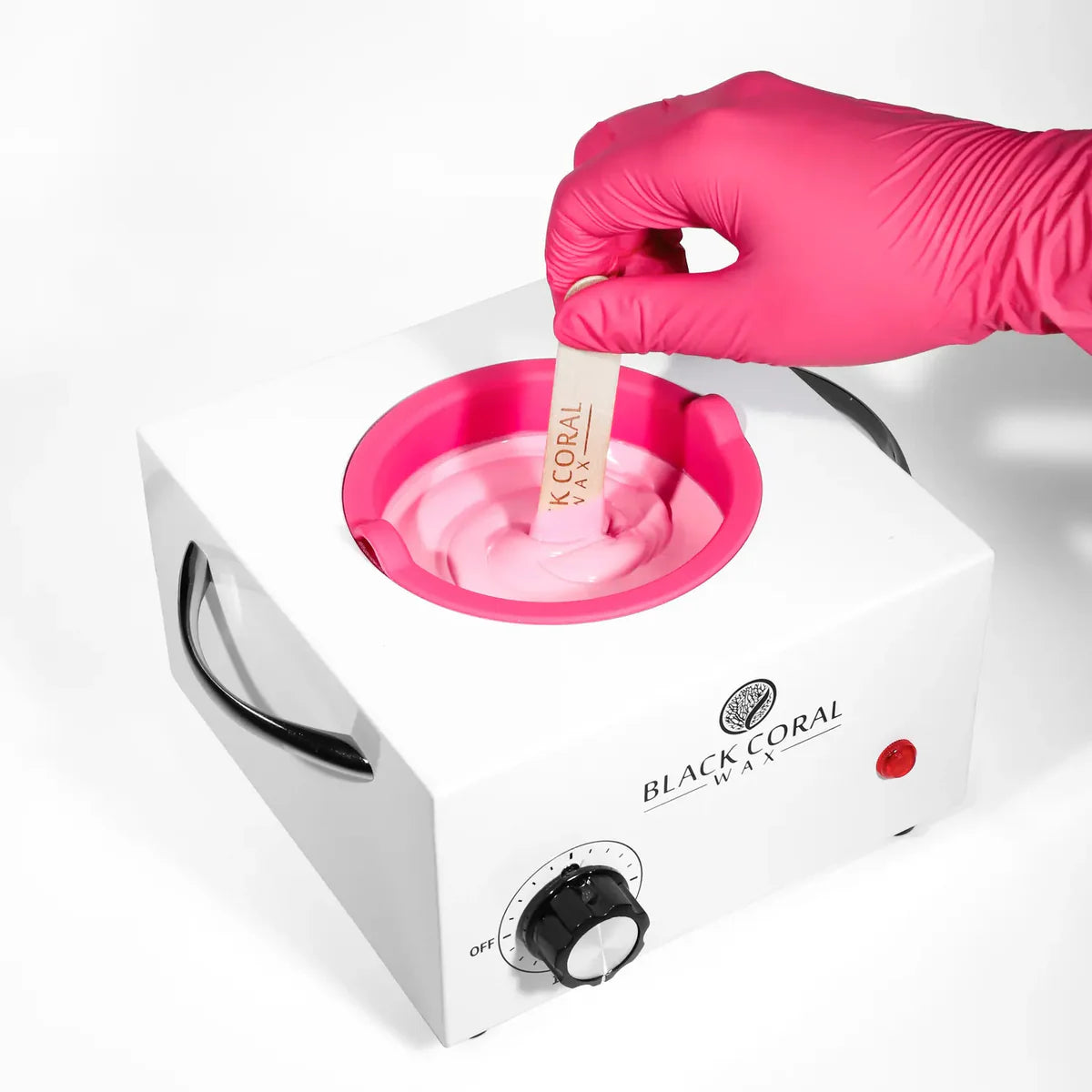A gloved hand dips a wooden spatula into melted pink wax in the Black Coral Wax USA Single (1lb) Wax Warmer, ready for hair removal against a white background.