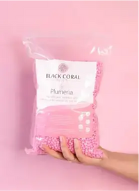A hand holding a large, clear plastic bag filled with small pink beads against a pink background. The bag is labeled Black Coral Hawaii and Plumeria.